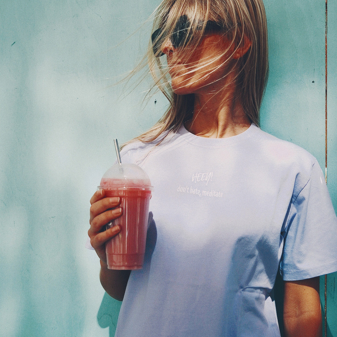 Heey! Plant Protein, maku Strawberry; tuote on kylmä vaaleanpunainen smoothie kirkkaassa muovimukissa litteine kansineen ja pillillä; sitä pitelevällä naisella tuulessa liehuvat vaaleat hiukset ja aurinkolasit; hänellä vaaleanvioletti mattapintainen puuvillapaita mustin lihavoiduin tekstein: HEEY! don't hate, meditate; tausta sileä yhtenäinen sininen seinä; kuva vyötäröstä ylöspäin ulkona.