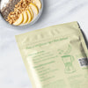Heey! Plant Protein, maku Vanilla-Pear; granola- ja hedelmäjogurttikulho keltaisen ruokapussin vieressä, jossa smoothie-reseptejä ja tehosekoittimen kuvitus, kaikki aseteltuna vaalealle marmoripinnalle; ruokapussi kiiltäväpintaista materiaalia painetuin grafiikoin, suorakaiteen muotoinen, lihavoitu groteskiteksti; ei ihmisiä tai käsiä; yläviistosta kuvattu tuotekuva.