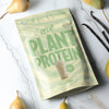 Heey! Plant Protein, maku Vanilla-Pear; beige-vihreä pussi tekstillä PLANT PROTEIN Vanilla Pear, kuva smoothiesta, marmoripinnalla tuoreiden päärynöiden ympäröimänä, korostaen kasvipohjaista proteiiniseosta; pakkaus mattapintainen, suorakaiteen muotoinen ja pystyssä; etiketissä lihavoitu groteskiteksti tuotenimelle; ei ihmisiä tai käsiä kuvassa.