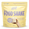 Heey! Vegan Food Shake, maku Vanilla; keltainen mattapintainen pussi tekstillä Heey! Food Shake pystyssä tasaisella alustalla; lihavoitua valkoista ja mustaa groteskitypografiaa, etiketissä complete meals, 25 vitamins & minerals ja 33g protein; ihmiskäsi pitelee kirkasta lasillista vaaleaa vanilja-shaketta pussin vieressä; tausta ei näytä yksityiskohtia.