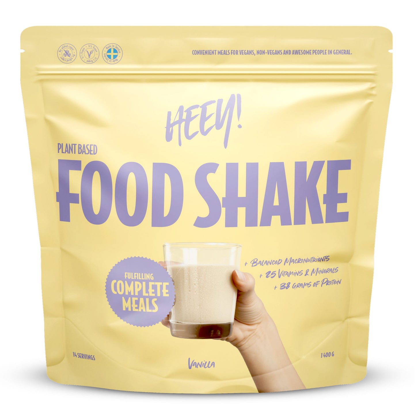 Heey! Vegan Food Shake, maku Vanilla; keltainen mattapintainen pussi tekstillä Heey! Food Shake pystyssä tasaisella alustalla; lihavoitua valkoista ja mustaa groteskitypografiaa, etiketissä complete meals, 25 vitamins & minerals ja 33g protein; ihmiskäsi pitelee kirkasta lasillista vaaleaa vanilja-shaketta pussin vieressä; tausta ei näytä yksityiskohtia.