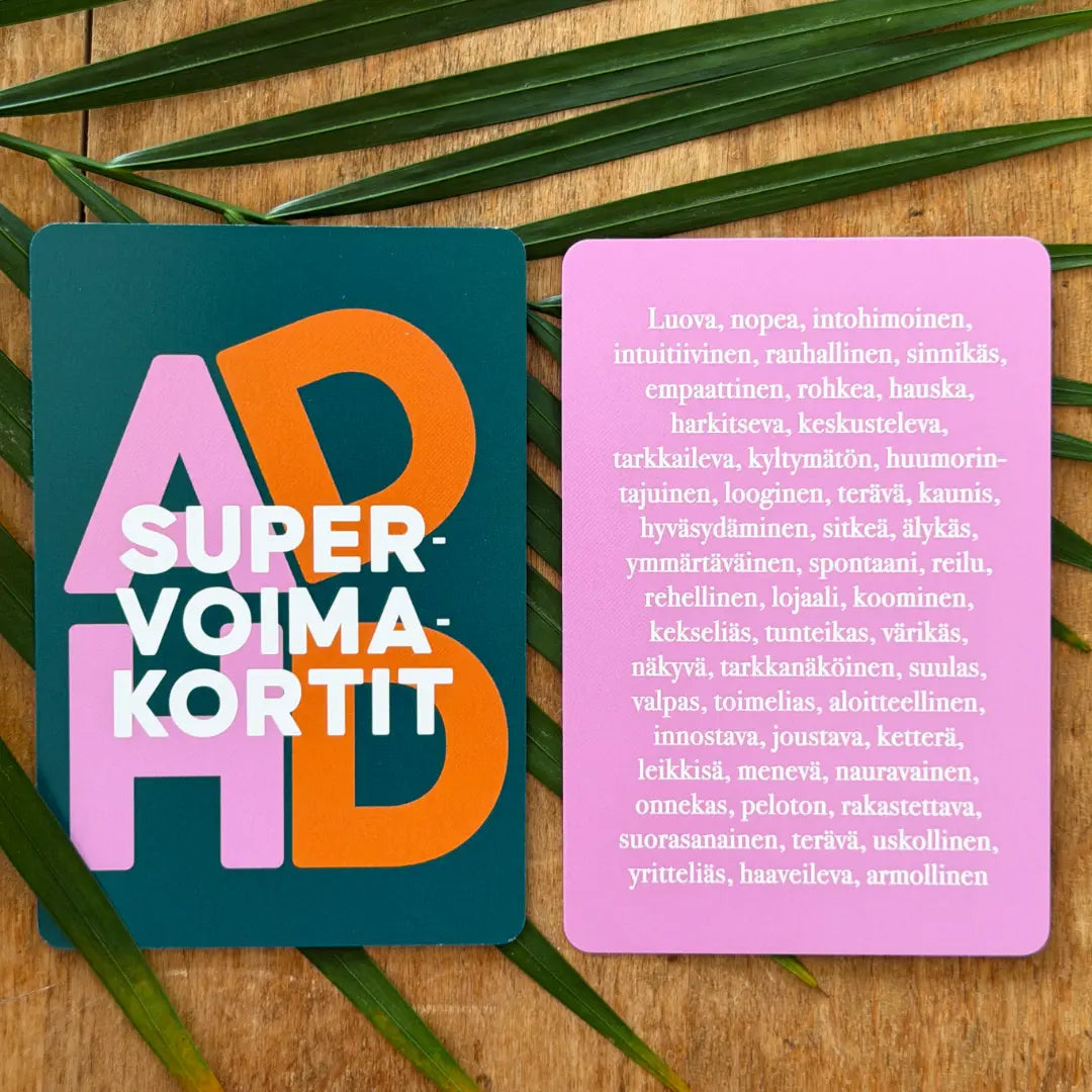 Hidasta Elämää ADHD-supervoimakortit: kaksi värikästä korttia puupinnalla vihreiden lehtien keskellä; toisessa lukee "ADHD SUPERVOIMAKORTIT", toisessa luetellaan positiivisia piirteitä suomeksi eloisalla pinkillä pohjalla; molemmat kortit ovat suorakulmaisia, painettu kortistolle mattapinnalla, lihavoitu groteskikirjoitus; ei ihmisiä tai käsiä näkyvissä; kuva on otettu ylhäältä ja rajattu tiiviisti tuotteisiin.
