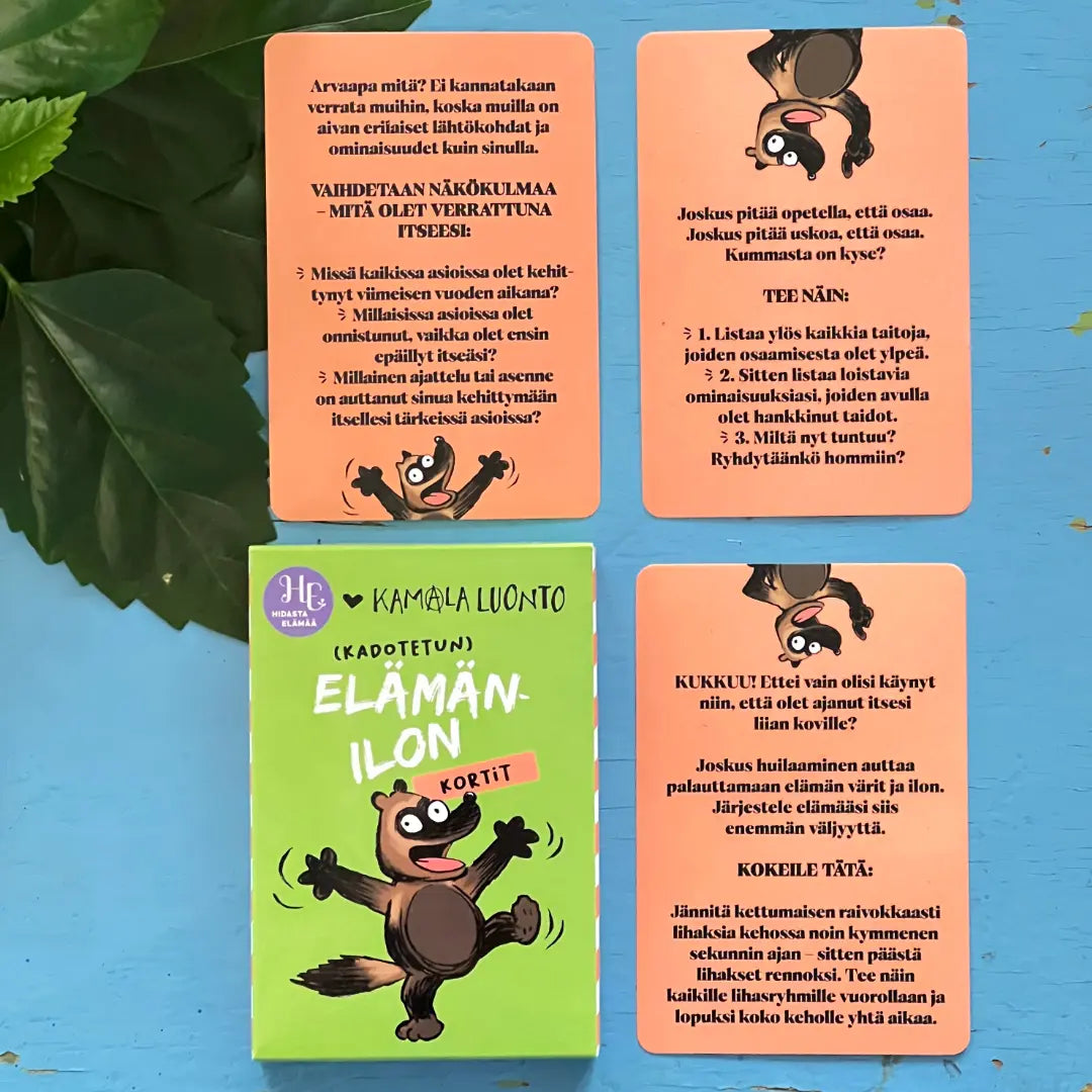 Hidasta Elämää Elämänilon kortit; neljä kuvitettua suomenkielistä peliä korttia, joissa jokaisessa piirretty orava, aseteltuna vihreän mattapintaisen suorakaiteen muotoisen korttilaatikon Elämänilon kortit ympärille lihavoidulla groteskifontilla; kaikki esineet sinisellä puisella pinnalla, jossa näkyvä syykuvio; muutama vihreä lehti yhdessä kulmassa; ei ihmisiä tai käsiä; kuvattu yläviistosta.
