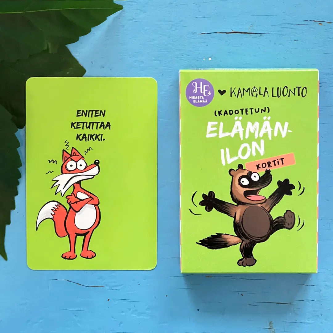 Hidasta Elämää Elämänilon kortit; vihreä kortti piirretyllä ketulla seisoo iloista piirrettyä majavaa esittävän vihreän laatikon vieressä; molemmat valmistettu paperista tai kartongista mattapinnalla; kortissa ja laatikossa pyöristetyt kulmat sekä leikkisää, lihavoitua suomenkielistä tekstiä: kortissa lukee Eniten ktaa kaikki ja laatikossa Elämän-ilon kortit; tuotteet neutraalilla taustalla hyvin valaistussa studiokuvassa.