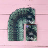 A set of dark green floral cards is fanned out on a pink wooden surface. The cards have a rectangular shape and feature matte finishes. One card is separated, displaying the text Hetken VOIMA-KORTIT in metallic gold serif letters with thin white botanical illustrations. No packaging, people, or actions are visible in the image.