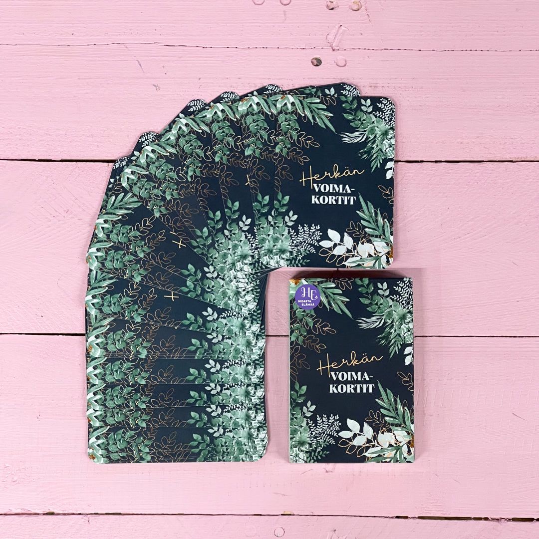 A set of dark green floral cards is fanned out on a pink wooden surface. The cards have a rectangular shape and feature matte finishes. One card is separated, displaying the text Hetken VOIMA-KORTIT in metallic gold serif letters with thin white botanical illustrations. No packaging, people, or actions are visible in the image.