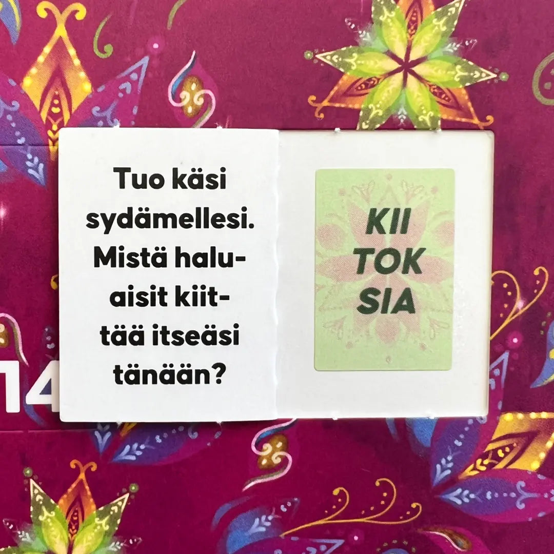 Hidasta Elämää Onnenhetkien Joulukalenteri; avattu kalenteriluukku paljastaa suomenkielistä tekstiä valkoisilla ja vihreillä suorakaiteen muotoisilla korteilla; kortit lihavoidulla mustalla groteskitekstillä, mattapinta, tiiviisti aseteltuina; tausta punais-violetti kukkapinta; ei ihmisiä tai käsiä kuvassa.