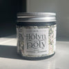 Hölynpöly Talivoide Natural: pieni pyöreä purkki tekstillä "Hölyn pöly Natural Ihovoide - Hudkräm" ja kukkakuvitus mattapintaisessa paperietiketissä, aseteltuna sileälle valkoiselle pinnalle; etiketin kirjasin on sekakokoista mustaa tekstiä; purkin kansi on metallinvärinen ja heijastava; auringonvalo luo pehmeitä varjoja ja korostaa etiketin yksityiskohtia; ei ihmisiä tai käsiä näkyvissä.