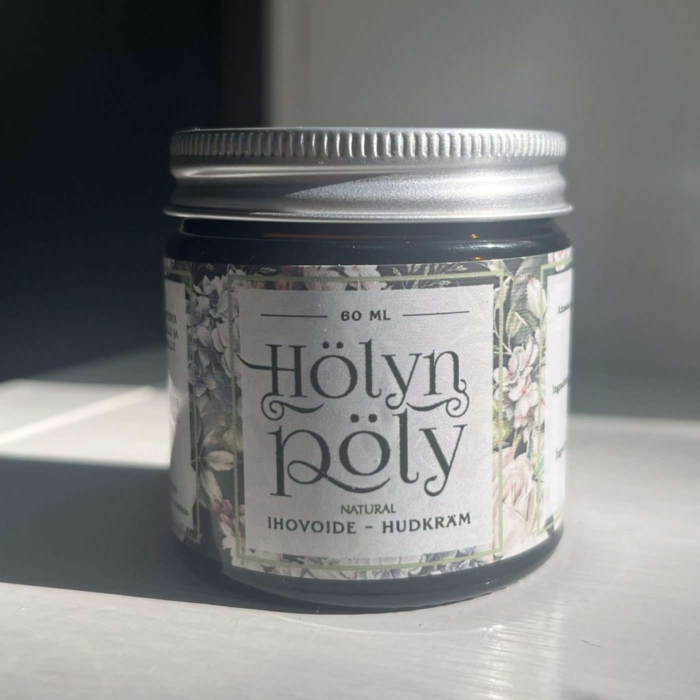 Hölynpöly Talivoide Natural: pieni pyöreä purkki tekstillä "Hölyn pöly Natural Ihovoide - Hudkräm" ja kukkakuvitus mattapintaisessa paperietiketissä, aseteltuna sileälle valkoiselle pinnalle; etiketin kirjasin on sekakokoista mustaa tekstiä; purkin kansi on metallinvärinen ja heijastava; auringonvalo luo pehmeitä varjoja ja korostaa etiketin yksityiskohtia; ei ihmisiä tai käsiä näkyvissä.