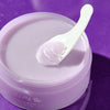 Hwarang Bellflower Cleansing Balm; valkoinen lasta pitelee kauhallista sileää, pastellivioletin väristä voidetta avoimen purkin päällä yhteensopivaa violettia taustaa vasten, korostaen tuotteen silkkistä tekstuuria; voide säilytetty pyöreässä purkissa ilman näkyvää brändäystä tai etikettitekstiä; sekä lastan että kannen pinta mattapintainen; ei ihmisiä tai käsiä; kuva rajattu tiiviisti ja otettu ylhäältä.