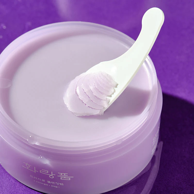Hwarang Bellflower Cleansing Balm; valkoinen lasta pitelee kauhallista sileää, pastellivioletin väristä voidetta avoimen purkin päällä yhteensopivaa violettia taustaa vasten, korostaen tuotteen silkkistä tekstuuria; voide säilytetty pyöreässä purkissa ilman näkyvää brändäystä tai etikettitekstiä; sekä lastan että kannen pinta mattapintainen; ei ihmisiä tai käsiä; kuva rajattu tiiviisti ja otettu ylhäältä.