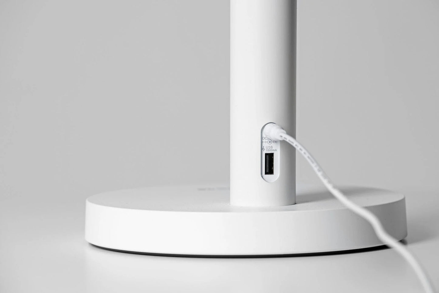 Innolux Oslo LED Kirkasvalo Pöytävalaisin; lähikuva modernin valkoisen lampun jalustasta, jossa sisäänrakennettu USB-portti; jalusta sileä ja mattapintainen, lieriömäinen muoto; valkoinen USB-kaapeli kytketty suorakaiteen muotoiseen porttiin; ei näkyviä etikettejä tai merkintöjä lampussa tai kaapelissa; tausta yksinkertainen ja vaaleanharmaa, ei muita esineitä näkyvissä.