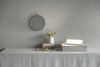A table with a gray tablecloth holds a vase of flowers, books, a tray with a bright desk lamp, and small decor items. The lamp is metallic with a dome-shaped shade and glossy finish. The books have plain covers without visible text. The tray is rectangular and made of light wood. A round mirror hangs on the gray textured wall above the table.