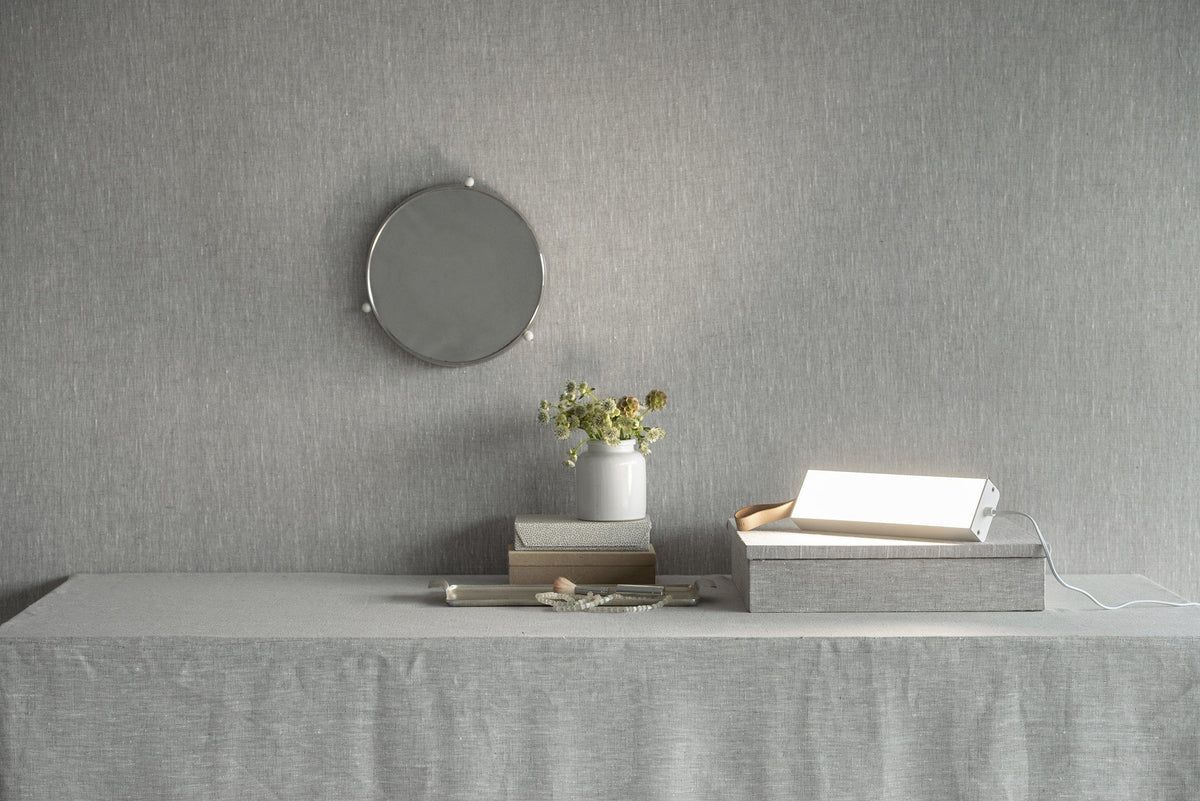 A table with a gray tablecloth holds a vase of flowers, books, a tray with a bright desk lamp, and small decor items. The lamp is metallic with a dome-shaped shade and glossy finish. The books have plain covers without visible text. The tray is rectangular and made of light wood. A round mirror hangs on the gray textured wall above the table.