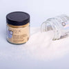JOIK Organic Warming Magnesium Jalkakylpysuola; kaksi lasipurkkia, merkitty Warming Ginger & Mustard Magnesium Footbath-Salt, toinen pystyssä ja toinen kaatuneena, näyttävät rauhoittavaa magnesiumkylpysuolaa valuneena valkoiselle pinnalle; kirkkaat sylinterinmuotoiset lasiastiat mustin kierrekorkein; sisältö on karkeaa murretun valkoista kidettä; etiketeissä pelkistettyä mustaa tekstiä mattapintaisella valkoisella paperilla; tausta puhdas ja yksivärinen valkoinen; ei ihmisiä tai käsiä näkyvissä.