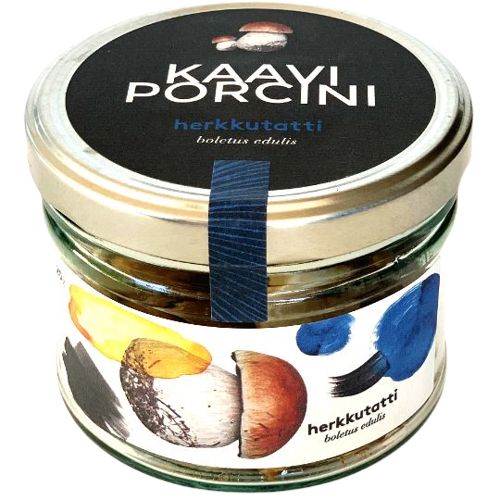 A glass jar with a metal lid labeled KAAYI PORCINI herkkutatti boletus edulis, featuring colorful mushroom illustrations and blue accents on the gourmet packaging. The cylindrical jar is clear glass, the label has bold uppercase lettering, and the surface is glossy. Background is neutral. No people or hands are present.