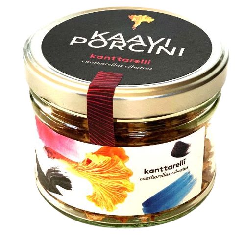 A glass jar of Kaayni brand dried chanterelle mushrooms, labeled kanttarelli cantharellus cibarius. The jar is cylindrical and transparent with a glossy black lid bordered by a silver metallic rim. The label has yellow, blue, and black brushstrokes on white and includes product names in clear sans-serif font. Studio setting with a plain background.