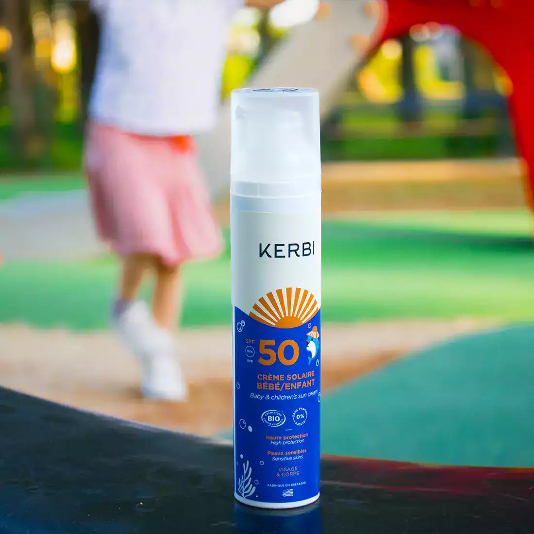 Kerbi Aurinkovoide Lapsille SPF 50; valkoinen muovipullo tekstillä ”Kerbi SPF 50 sunscreen for babies and children” pystyssä monivärisellä alustalla ulkona; pullo kiiltäväpintainen yksinkertaisella, lihavoidulla typografialla etiketissä; sumeana taustalla leikkivä lapsi vaaleanpunaisessa hameessa.