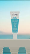 Kerbi Kosteuttava Kasvovoide SPF 30; tuubi KERBI Crème Solaire Hydratante SPF 30 -aurinkovoidetta seisoo pystyssä valkoisella kaidepinnalla, taustalla sumea meri ja pastellinvärinen taivas; tuubi lieriömäinen, pystyssä, muovia mattapinnalla; etikettitekstissä tuotenimi ja SPF-taso selkeällä groteskilla; ei ihmisiä tai käsiä näkyvissä; tuotekuva ulkoilmamaisemaa vasten.