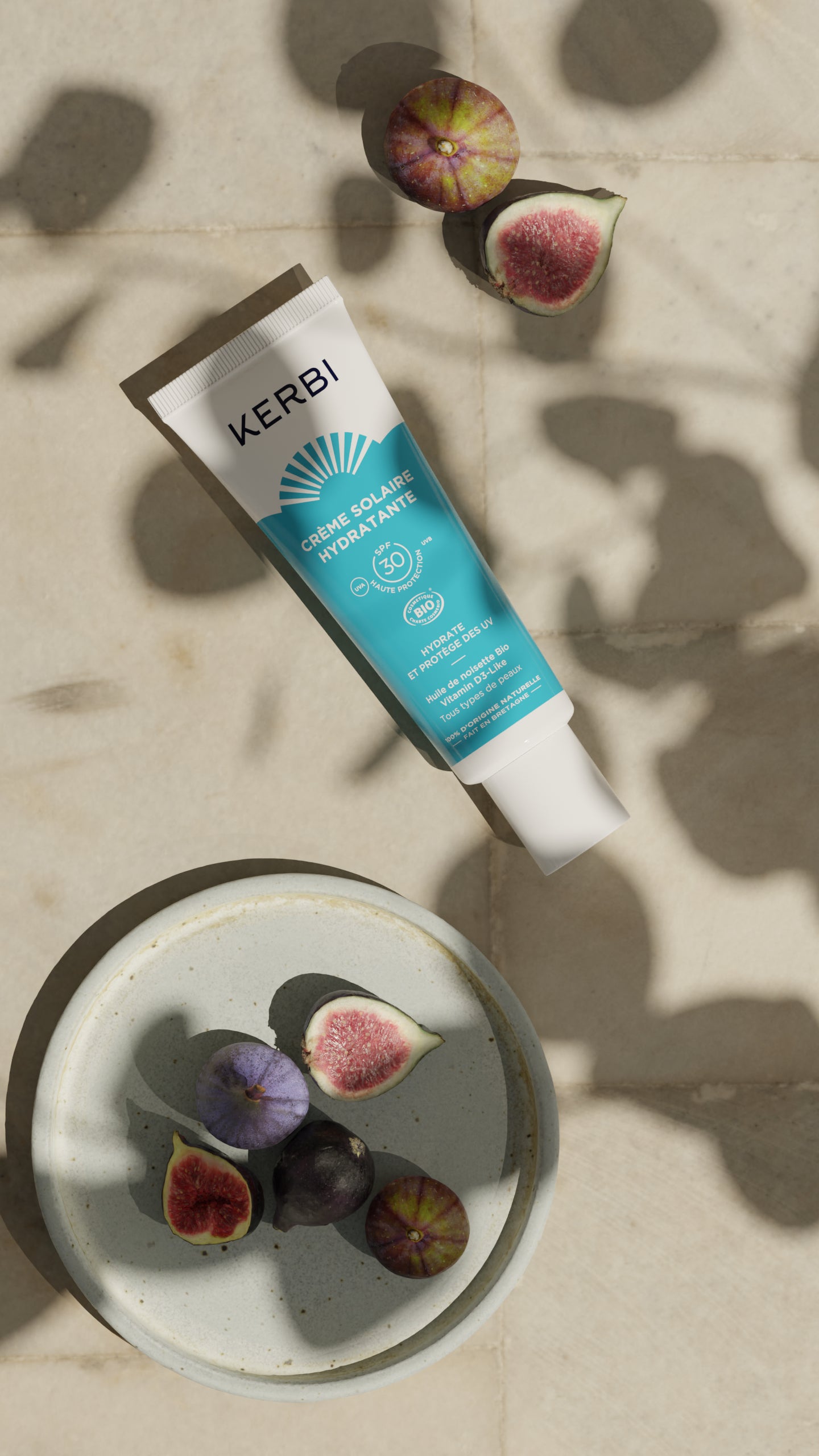 Kerbi Kosteuttava Kasvovoide SPF 30; tuubi Kerbi-aurinkovoidetta mattapinnalla, vaalea pakkaus ja groteskietikettiteksti, aseteltuna vaalealle tasaiselle alustalle; vieressä pieni kulho ja kaksi puolikasta viikunaa; kuvassa selkeitä lehtien ja viikunoiden varjoja; ei ihmisiä tai käsiä näkyvissä.