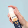 Kerbi Aurinkovoide SPF 30; käsi pitelee KERBI SPF 30 Sun Lotion -pulloa vaaleansinistä taustaa vasten, pumppuannostelija hieman painettuna ja aurinkovoidetta näkyvissä; pullo valkoista muovia puolikiiltävällä pinnalla, lieriömäinen muoto ja oranssi pumppumekanismi; etiketissä lihavoitua mustaa groteskitekstiä ”KERBI” ja selkeää oranssia tekstiä ”SPF 30”.