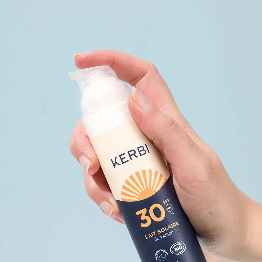 Kerbi Aurinkovoide SPF 30; käsi pitelee KERBI SPF 30 Sun Lotion -pulloa vaaleansinistä taustaa vasten, pumppuannostelija hieman painettuna ja aurinkovoidetta näkyvissä; pullo valkoista muovia puolikiiltävällä pinnalla, lieriömäinen muoto ja oranssi pumppumekanismi; etiketissä lihavoitua mustaa groteskitekstiä ”KERBI” ja selkeää oranssia tekstiä ”SPF 30”.