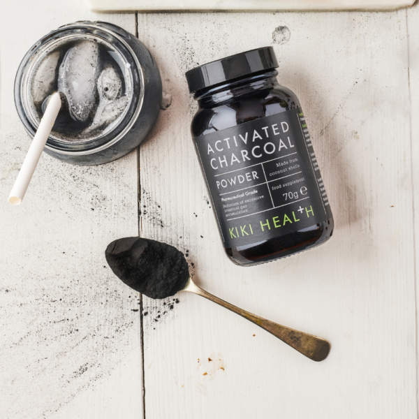 Kiki Health Aktiivihiilijauhe; purkki tekstillä ”Activated Charcoal Powder” lepää valkoisella pinnalla metallilusikan vieressä, jossa löysää, mattamustaa jauhetta; lähellä avoin muoviastia, jossa valkoinen tikku sisällä; etiketissä lihavoitua, selkeää tekstiä; mustaa jauhetta siroteltuna sileälle valkoiselle taustalle; lähikuva tuotteesta, ei ihmisiä näkyvissä.