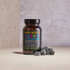 Kiki Health Body Biotics Gummies Maitohappobakteeri; purkki, merkitty Body Biotics Gummies for Children by Kiki Health, seisoo beigellä pinnalla useiden tummien probioottipurukumien edessä; etiketissä groteskitypografiaa valkoisena ja vihreänä mustaa taustaa vasten; purkki on sylinterinmuotoinen, tummanruskeaa läpikuultavaa muovia kiiltäväpinnalla ja mustalla kannella; tausta vaaleanharmaa ja tekstuuripintainen.