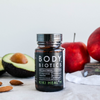 Kiki Health Body Biotics Maitohappobakteeri; pullo Kiki Health Body Biotics -lisäravinnetta seisoo pöydällä avokadon, manteleiden, punaisten omenien ja valkoisen liinan kera taustalla; tumma lasipullo on sylinterinmuotoinen kierrekorkilla, etiketissä mattamusta pohja ja yksinkertaista valkoista groteskitekstiä ”Kiki Health Body Biotics”; ei ihmisiä tai käsiä näkyvissä; laaja kuvasommittelu.