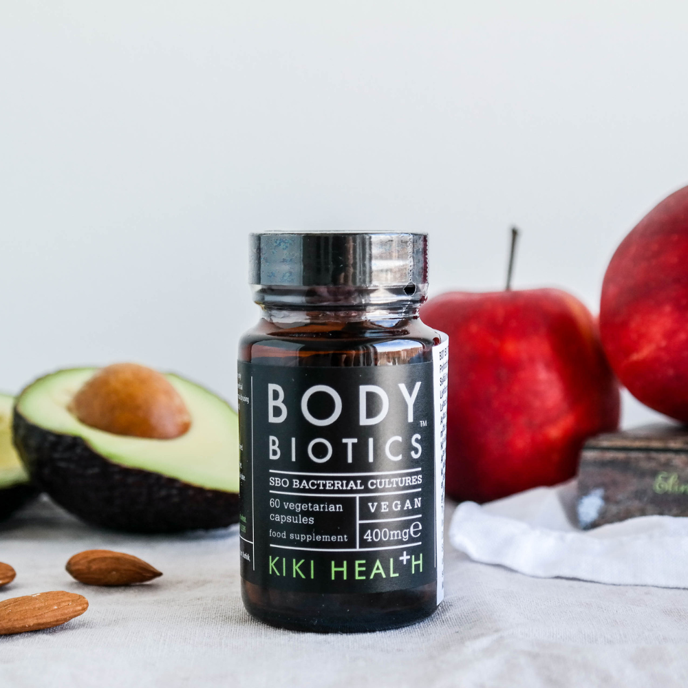 Kiki Health Body Biotics Maitohappobakteeri; pullo Kiki Health Body Biotics -lisäravinnetta seisoo pöydällä avokadon, manteleiden, punaisten omenien ja valkoisen liinan kera taustalla; tumma lasipullo on sylinterinmuotoinen kierrekorkilla, etiketissä mattamusta pohja ja yksinkertaista valkoista groteskitekstiä ”Kiki Health Body Biotics”; ei ihmisiä tai käsiä näkyvissä; laaja kuvasommittelu.