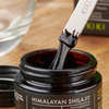 Kiki Health Himalayan Shilajit; lähikuva purkista tekstillä ”Himalayan Shilajit”, lasta nostaa paksua, tummaa hartsia esiin; purkki tummaa kiiltävää lasia pyöreässä muodossa mustalla kierrekannella; lasta ohut ja metallinen; etikettiteksti lihavoidulla tikkukirjasimella; taustalla tasaisella pinnalla sumeana tuoterasia ja kirkas lasikulho; ei ihmisiä näkyvissä kuvassa.