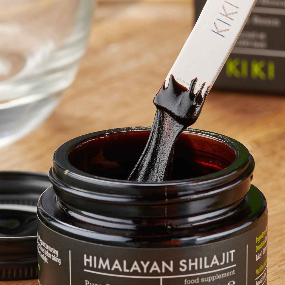 Kiki Health Himalayan Shilajit; lähikuva purkista tekstillä ”Himalayan Shilajit”, lasta nostaa paksua, tummaa hartsia esiin; purkki tummaa kiiltävää lasia pyöreässä muodossa mustalla kierrekannella; lasta ohut ja metallinen; etikettiteksti lihavoidulla tikkukirjasimella; taustalla tasaisella pinnalla sumeana tuoterasia ja kirkas lasikulho; ei ihmisiä näkyvissä kuvassa.
