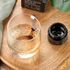 Kiki Health Himalayan Shilajit; vesilasi metallisen sekoituslusikan kera avoimen, pyöreän mustan lasipurkin vieressä, jossa teksti ”Himalayan Shilajit by Kiki Health” valkoisella tikkukirjasimella mattamustalla etiketillä; esineet sileällä, vaaleasävyisellä pinnalla; lähellä taiteltu vihreä liina ja yksittäinen litteä vihreä lehti; ei ihmisiä tai käsiä näkyvissä; laaja kuva ylhäältä.