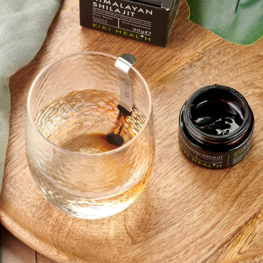 Kiki Health Himalayan Shilajit; vesilasi metallisen sekoituslusikan kera avoimen, pyöreän mustan lasipurkin vieressä, jossa teksti ”Himalayan Shilajit by Kiki Health” valkoisella tikkukirjasimella mattamustalla etiketillä; esineet sileällä, vaaleasävyisellä pinnalla; lähellä taiteltu vihreä liina ja yksittäinen litteä vihreä lehti; ei ihmisiä tai käsiä näkyvissä; laaja kuva ylhäältä.
