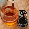 Kiki Health Himalayan Shilajit; lasi meripihkanväristä nestettä metallisen lusikan kera puisella alustalla avoimen purkin vieressä, jossa teksti ”Himalayan Shilajit” ja tummaa, paksua tahnaa sisällä; purkki musta kultaisin ja valkoisin antiikvatekstein; kuva hyvin valaistu, pöytäkuvakulma, ei ihmisiä tai käsiä näkyvissä; tausta tasainen ja neutraali.