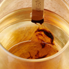 Kiki Health Himalayan Shilajit; lähikuva teelasista, jossa teepussi osittain upotettuna, tummien teevirtausten sekoittuessa kultaiseen nesteeseen; kirkas lasi lieriömäinen, sileä ja kiiltäväpintainen; ei näkyviä etikettejä tai pakkausta, ei brändäystä tai typografiaa; tausta ja ympäristö eivät näy tässä tiiviisti rajatussa kuvassa; ei käsiä tai ihmisiä näkyvissä.
