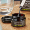 Kiki Health Himalayan Shilajit; purkki KIKI Health Himalayan Shilajit Pure Resin -tuotetta kiiltävällä mustalla kierrekannella, avoinna sileällä puupinnalla; purkki lieriömäinen, tumman lasin tai muovin väristä, tuotenimi lihavoidulla valkoisella ja vihreällä tekstillä mattamustalla etiketillä; metallinen lasta pitelee tummaa, sitkeää hartsia purkin yläpuolella; ei ihmisiä näkyvissä kuvassa.