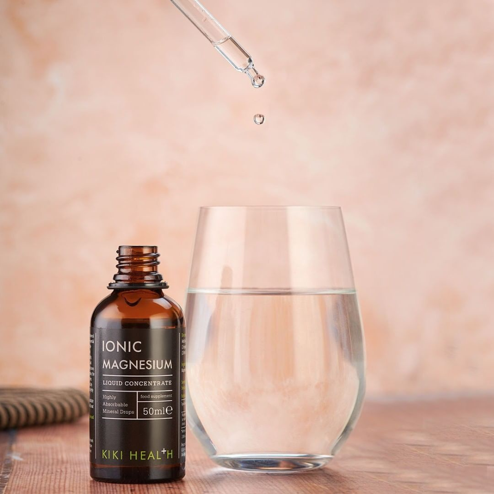 Kiki Health Ionisoitu Magnesium; tiputin annostelee nestettä vesilasin yläpuolelle ruskean pullon vieressä, merkitty Ionic Magnesium by KIKI Health, vaalealla pinnalla, korostaen ionisoidun magnesiumlisän käyttöä; pullo on sylinterinmuotoinen mattamustalla kierrekorkilla ja selkeällä valkoisella ja vihreällä groteskitekstillä; tausta yksivärinen, valaistus tasainen, ei ihmisiä tai käsiä kuvassa.