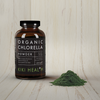 Kiki Health Luomu Chlorella-jauhe; ruskea lasipurkki valkoisella etiketillä, jossa lukee ”Organic Chlorella Powder”, seisoo pienen hienoksi jauhetun vihreän jauhekasan vieressä; purkki on sylinterinmuotoinen mattapinnalla; ympäristönä vaalea, hienosyinen puupinta pystysuuntaista puutekstuuria vasten; ei ihmisiä tai käsiä näkyvissä.