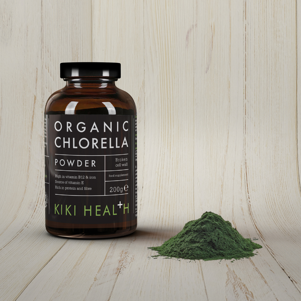 Kiki Health Luomu Chlorella-jauhe; ruskea lasipurkki valkoisella etiketillä, jossa lukee ”Organic Chlorella Powder”, seisoo pienen hienoksi jauhetun vihreän jauhekasan vieressä; purkki on sylinterinmuotoinen mattapinnalla; ympäristönä vaalea, hienosyinen puupinta pystysuuntaista puutekstuuria vasten; ei ihmisiä tai käsiä näkyvissä.