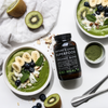 Kiki Health Natures Living Luomu Superfood-jauhe; kaksi smoothiekulhoa banaanin, mustikoiden, kiivin, manteleiden ja mintun kera lähellä kirkasta sylinterinmuotoista purkkia, jossa vihreä etiketti ”Natures Living Superfood Organic Blend” groteskifontilla, sekä pieni valkoinen kulho vihreää jauhetta; kaikki esineet mattavalkoisella pinnalla, kuvattu ylhäältä; ei ihmisiä tai käsiä näkyvissä.