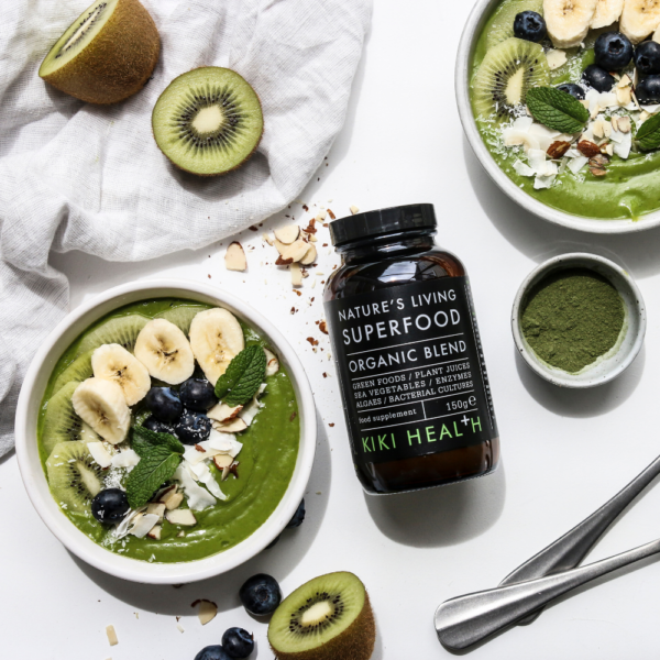 Kiki Health Natures Living Luomu Superfood-jauhe; kaksi smoothiekulhoa banaanin, mustikoiden, kiivin, manteleiden ja mintun kera lähellä kirkasta sylinterinmuotoista purkkia, jossa vihreä etiketti ”Natures Living Superfood Organic Blend” groteskifontilla, sekä pieni valkoinen kulho vihreää jauhetta; kaikki esineet mattavalkoisella pinnalla, kuvattu ylhäältä; ei ihmisiä tai käsiä näkyvissä.