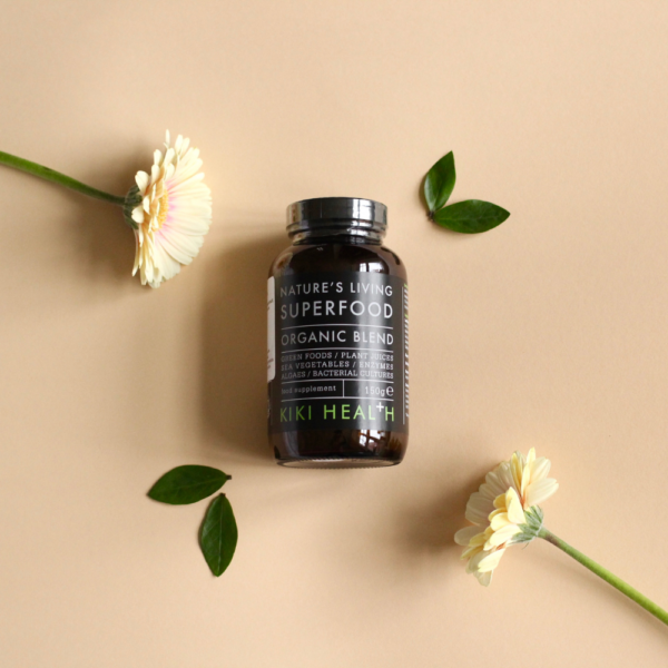 Kiki Health Natures Living Luomu Superfood-jauhe; purkki Kiki Health Natures Living Superfood Organic Blend -tuotetta on keskitetty mattapintaiselle beigelle alustalle; purkki on tumma, sylinterinmuotoinen ja lasia, mustalla kierrekorkilla; etiketissä valkoista groteskitekstiä ja vihreitä yksityiskohtia sekä näkyvissä oleva tuotenimi; koristeeksi aseteltuna kaksi vaaleankeltaista kukkaa ja useita vihreitä lehtiä; ei ihmisiä tai käsiä näkyvissä; kuva otettu ylhäältä.
