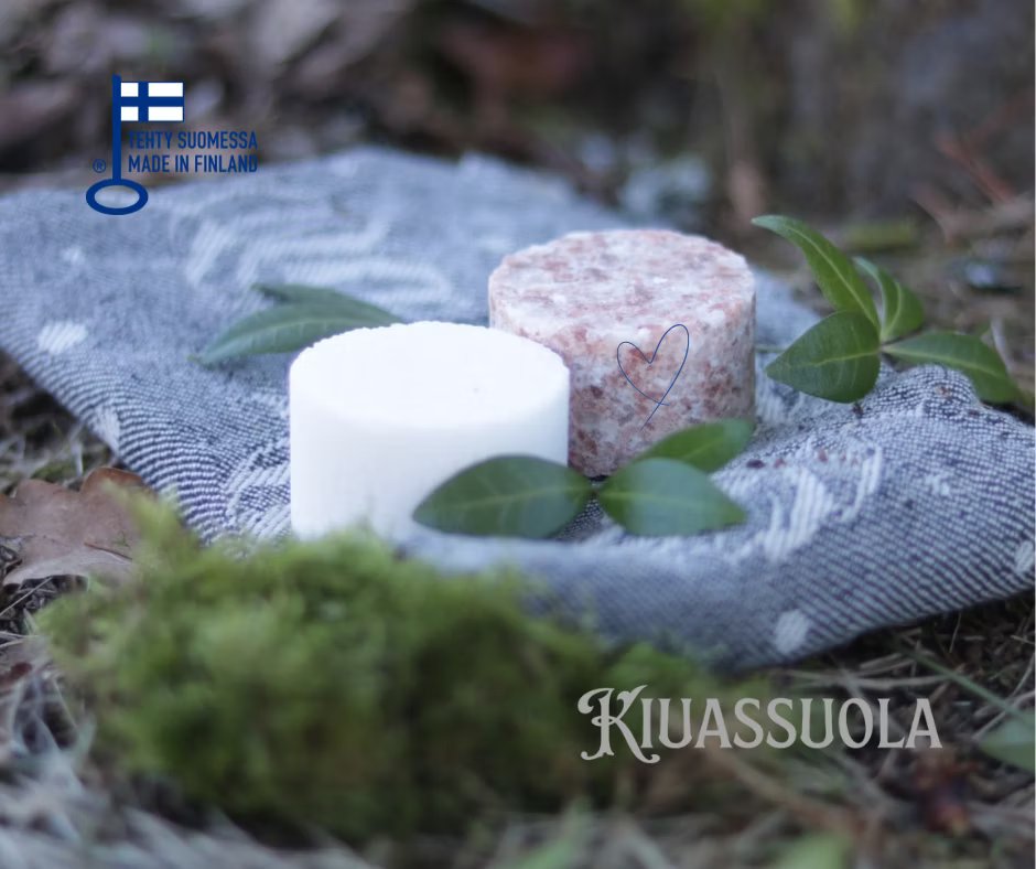 Kiuassuola; kaksi pyöreää saippuapalaa, toinen valkoinen ja toinen vaaleanpunainen, on aseteltu kuvioidulle harmaalle kankaalle, jossa näkyy vihreitä lehtiä, sammalta ja metsän pohjaa; molemmat kappaleet ovat mattapintaisia; pakkausta ei näy; teksti "Kiuassuola" ja "Made in Finland" selkeällä groteskilla suoraan kuvan päällä; ei ihmisiä tai käsiä näkyvissä.