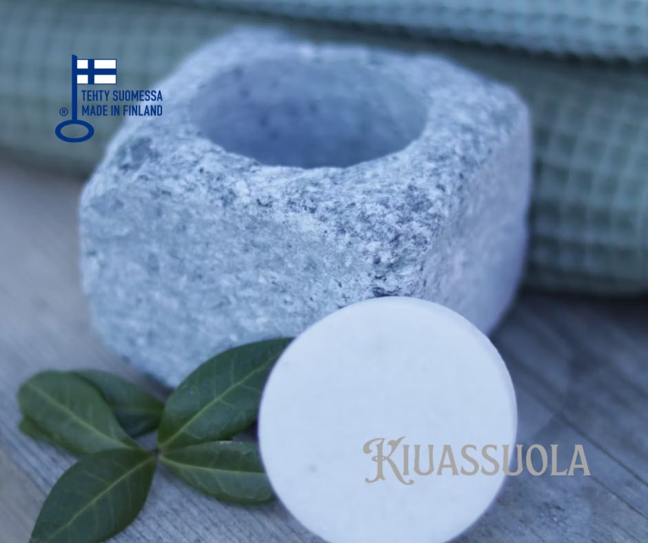 Kiuassuola ILMARI Aloituspakkaus; graniittinen suolapidike ja pyöreä valkoinen suolakiekko kahden vihreän lehden ja siististi taitetun vaalean pyyhkeen vieressä; suolapidike on mattapintainen ja pyöreä; kuvassa versaalilla groteskitekstillä "Made in Finland" ja "KIUASSUOLA"; tausta neutraali, ei ihmisiä tai käsiä.