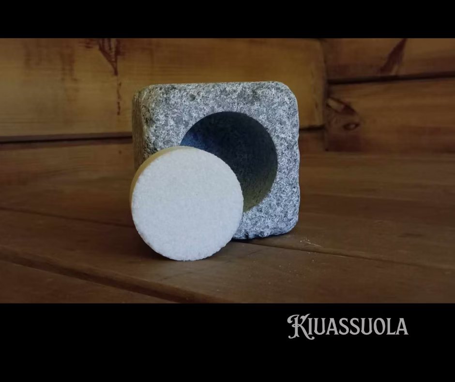 Kiuassuola ILMARI Aloituspakkaus; pyöreä valkoinen kiekko nojaa ontoksi koverrettuun harmaaseen kivikuutioon puupinnalla; kiekko on merkitsemätön, mattapintainen ja sileä; kivikuutio on tekstuuripintainen, siinä näkyvä syvennys ja terävät reunat; taustalla pystysuuntaista puupaneelia luonnonsävyissä; "KIUASSUOLA" groteskitekstillä kuvan oikeassa alakulmassa; ei ihmisiä tai pakkausta näkyvissä.