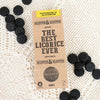Klepper & Klepper Gluteeniton Lakritsi Honey; suorakaiteen muotoinen pakkaus tekstillä The Best Licorice Ever on aseteltu valkoiselle pitsiliinalle, pyöreitä mustia lakritsipaloja ympärillä; pakkauksessa lihavoitua groteskitekstiä kertoen gluteenittomasta, liivatteettomasta ja hunajan makuisesta lakritsista; ei ihmisiä tai käsiä näkyvissä; kuva otettu yläviistosta.