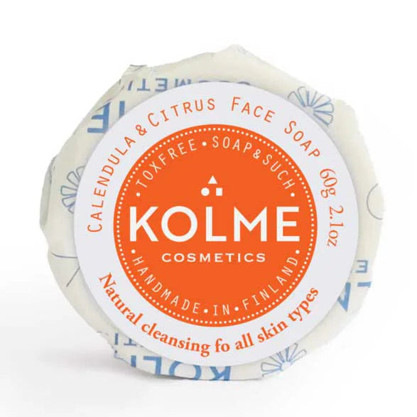 Kolme Cosmetics Kasvosaippua Kehäkukka & Sitrus: käärittyä saippuapalaa oranssi-valkoisella mattapaperietiketillä; palanen on suorakulmainen; etiketissä yksinkertainen groteskikirjasin mustalla ja oranssilla, pääteksti kertoo luonnollisesta puhdistuksesta kaikille ihotyypeille; tämä käsintehty, myrkytön saippua on valmistettu Suomessa; ei käsiä tai ihmisiä näkyvissä, kuvattu neutraalia taustaa vasten.