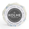 Kolme Cosmetics Kasvosaippua Teepuuöljy & Aktiivihiili: pyöreä saippuapalanen käärittynä valkoiseen paperiin sinisin kuvioin; ympyränmuotoisessa etiketissä groteskikirjasin ja teksti: "KOLME COSMETICS Teatree & Charcoal Face Soap"; käsintehty Suomessa yhdistelmä- ja epäpuhtaalle iholle, ainutlaatuinen kaava hyödyntää aktiivihiilen puhdistavia ominaisuuksia; tuote kuvattu yksinään neutraalia taustaa vasten, ei ihmisiä tai käsiä näkyvissä.