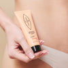 LIP Intiimialueen Hoitobalsami Macadamia + Kaura; henkilö pitelee valkoista tuubia, jossa lukee "LP Organic Intimate Care Lip Balm with macadamia oil", toisessa kädessä lantion lähellä, yllään beige alushousut; tuubissa on mustaa groteskitekstiä ja yksinkertainen kuvio mattapinnalla; tausta neutraali ja siisti; kuva rajattu vartaloon ja tuotteeseen.